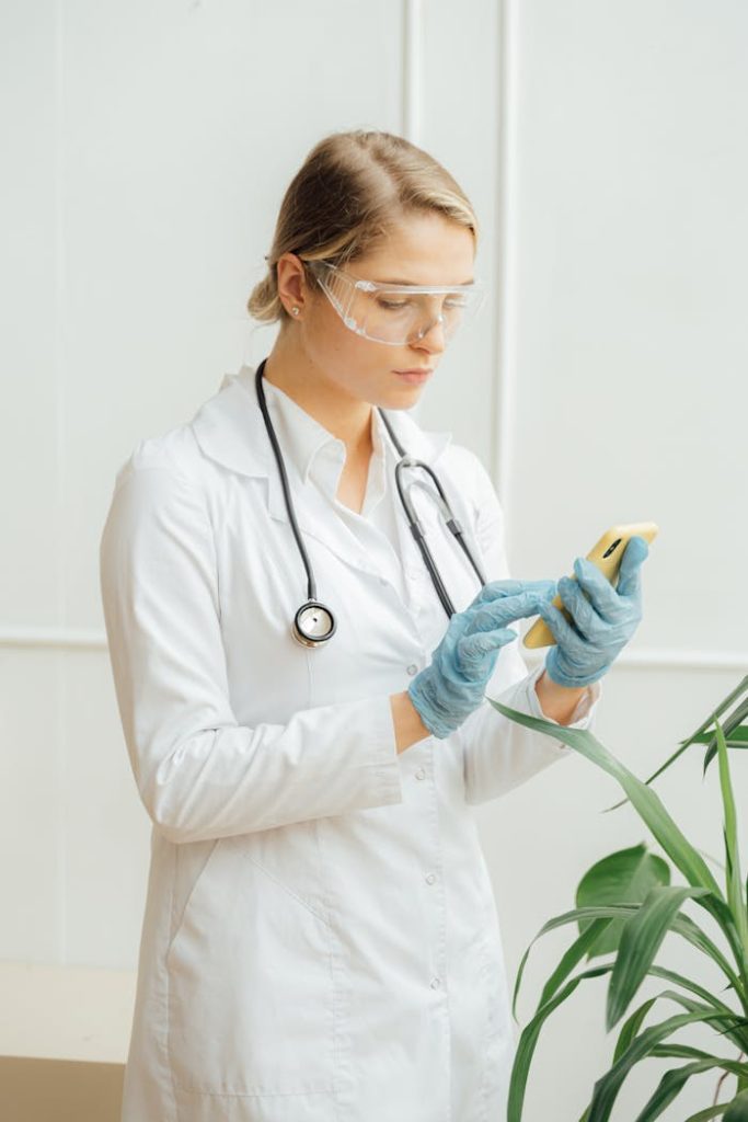 a-doctor-using-her-smartphone-5214960 A female doctor wearing protective gear and a white coat checks her smartphone in a medical office.